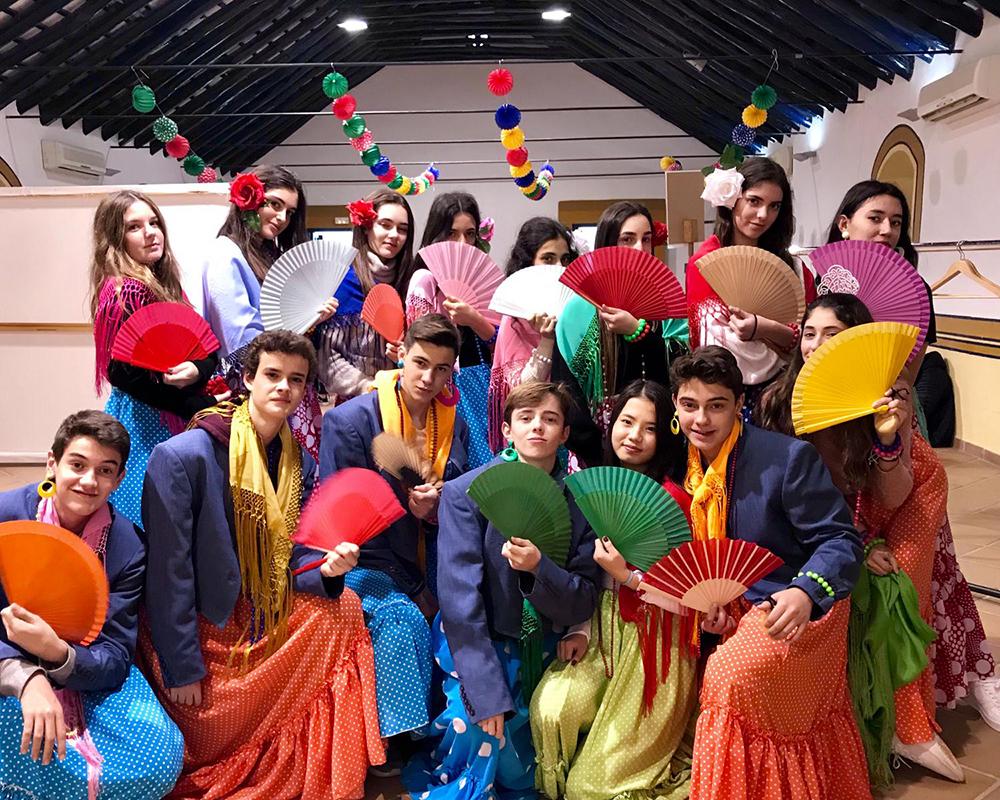 A group of students dressed in colorful flamenco costumes with fans, celebrating Spanish culture, in an indoor setting with decorations, showcasing cultural diversity and eng年龄ment.